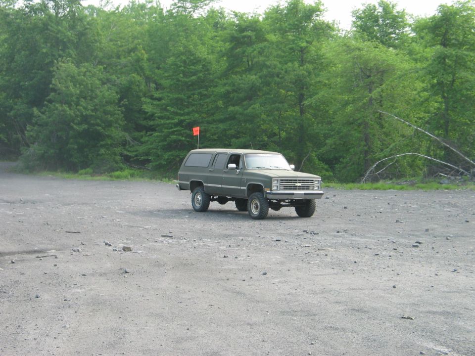 Olive drab square-body Chevrolet Suburban 4x4 with orange whip flag on a gravel lot