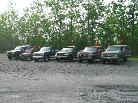 1995 Ford Bronco - A row of six 4x4s lined up front-first on a gray shale staging lot at a wooded off-road park, each flying an orange trail whip flag. The li…
