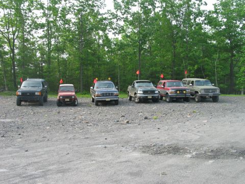 1995 Ford Bronco - A lineup of six off-road vehicles, including a 1995 Ford Bronco, are parked in a gravel lot with trees in the background.