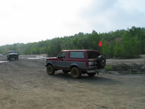 1995 Ford Bronco - A two-tone red and grey 1995 Ford Bronco is seen from the rear on a dirt road, with another vehicle in the distance.