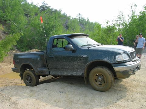 1995 Ford Bronco - Ford F-150 pickup truck navigates a muddy off-road course with trees and rocky terrain in the background.