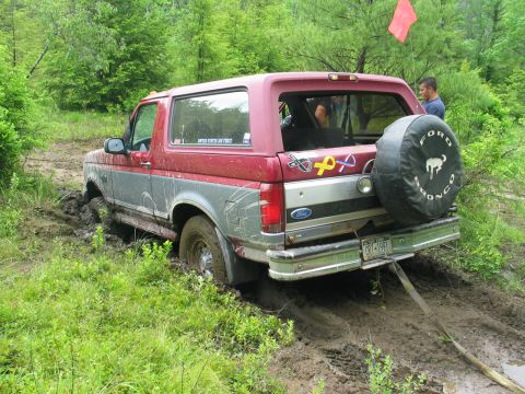 1995 Ford Bronco - Two-tone maroon-over-silver full-size Ford Bronco buried to the axles in a mud hole, a recovery strap running from the rear bumper out of f…