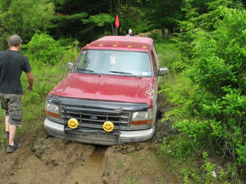 1995 Ford Bronco - Front view of a maroon full-size Ford Bronco sitting low in a deep muddy rut on a green wooded trail, mud spray up the hood and fender. It…