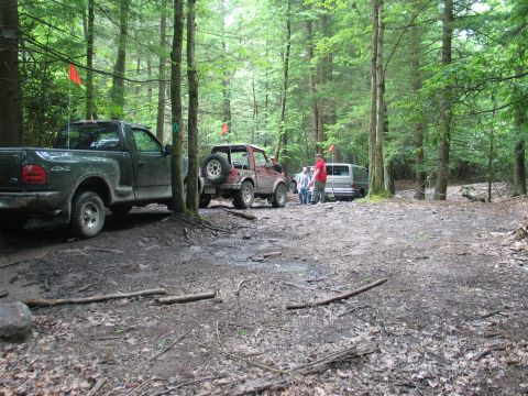 1995 Ford Bronco - A muddy trail staging area in a dense pine forest with a dark green Ford F-150 regular-cab 4x4 parked at left, a red soft-top Suzuki Samura…