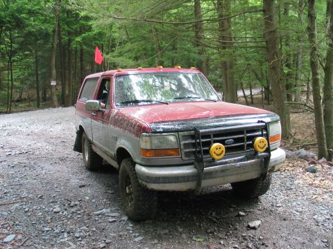 1995 Ford Bronco - Three-quarter front view of a mud-splattered red and silver two-tone Ford Bronco on a gravel trail in dense woods. It wears a black tube br…