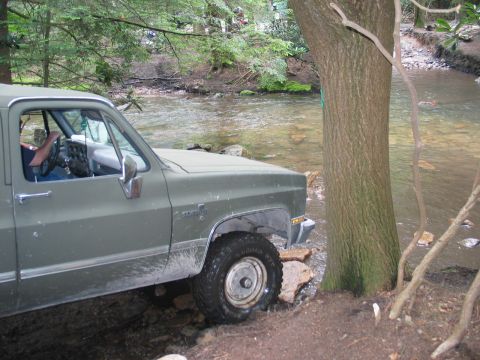 1995 Ford Bronco - A flat olive-green square-body Chevrolet Silverado pickup noses down a rocky bank into a shallow creek, shot from the driver's side with th…