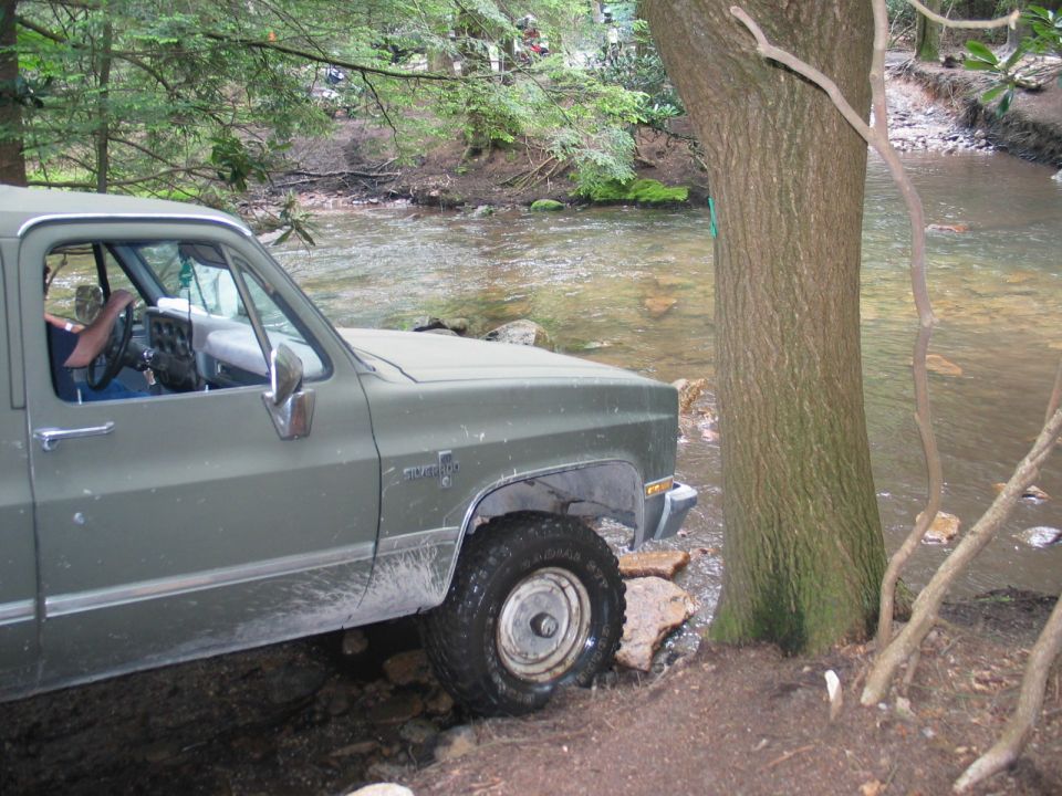 Olive green square-body Chevy Silverado pickup entering a shallow creek in the woods