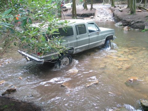 1995 Ford Bronco - A silver-grey square-body Chevrolet crew cab pickup fords a shallow rocky creek in the woods, water spraying off the rear tire and dripping…