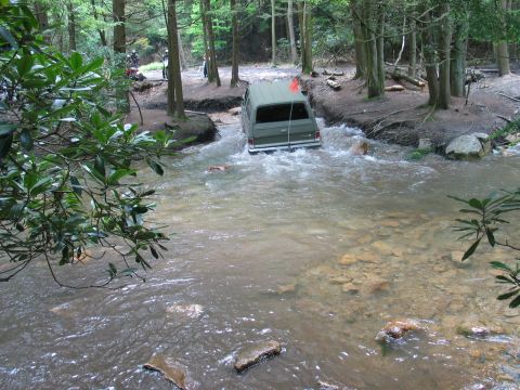 1995 Ford Bronco - Rear view of an olive-drab full-size SUV fording a wide, muddy creek in the woods, water pushing up around the rear bumper and tailgate. An…