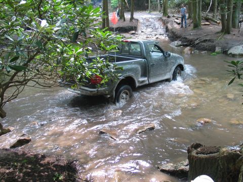 1995 Ford Bronco - Silver regular-cab Ford pickup fording a muddy rocky creek in a wooded trail park, water pushing up over the wheels as it drives away from…