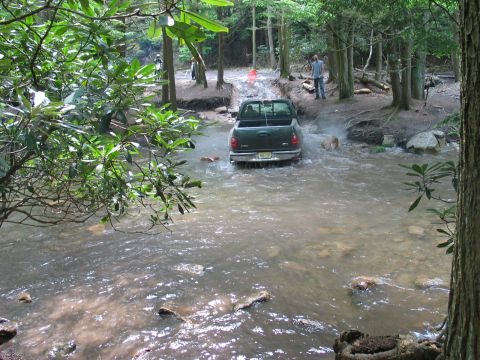 1995 Ford Bronco - Dark green Ford F-150 pickup fording a shallow rocky creek on a wooded trail, viewed from behind with water pushing up around the doors and…
