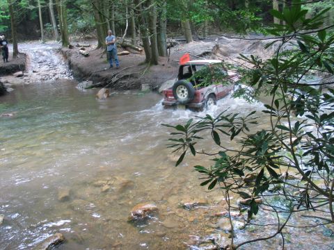 1995 Ford Bronco - Red full-size Bronco with a white removable top fords a shallow rocky creek in a hemlock forest, throwing spray off the front tires. A tail…