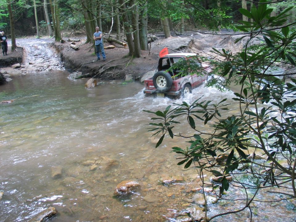 Red Bronco with white top fording a rocky forest creek, spare tire on tailgate