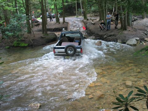 1995 Ford Bronco - Rear view of a topless black-and-silver Ford Bronco fording a fast-moving rocky creek, water breaking in white foam against the tailgate an…