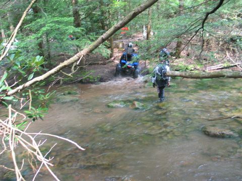 1995 Ford Bronco - A blue quad ATV fords a shallow, rocky forest creek while a rider in full gear and a backpack wades through the water alongside it.