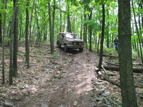 1995 Ford Bronco - Front three-quarter view of a mud-caked red 1990s Ford full-size SUV cresting a rocky wooded trail, fitted with a brush guard carrying yell…