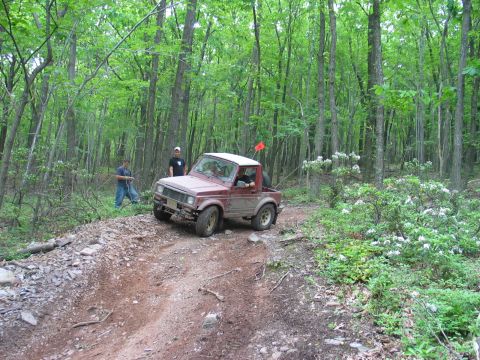 1995 Ford Bronco - A small maroon Suzuki Samurai-style mini 4x4 with a white hardtop, brush guard and orange whip flag sits on a rutted dirt trail cut through…