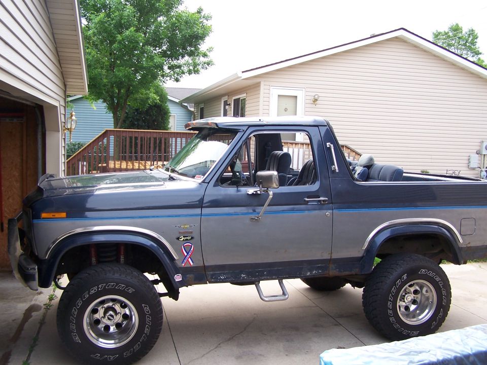 Gray and blue 1980s Ford Bronco lifted on chrome wheels, top off, in driveway