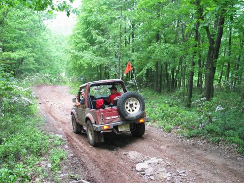 1995 Ford Bronco - Mud-splattered dark red Suzuki Samurai with the top removed climbing a wet dirt forest trail, carrying two occupants, a tailgate-mounted sp…