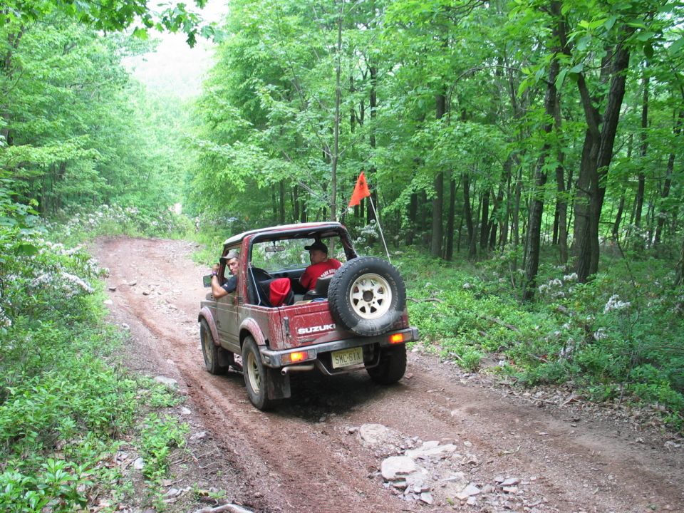 Muddy red topless Suzuki Samurai with whip flag on a wet dirt forest trail