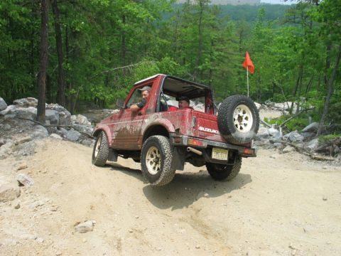 1995 Ford Bronco - Rear three-quarter view of a red soft-top Suzuki Samurai climbing a dirt-and-rock trail with two occupants aboard, mud splattered up the bo…