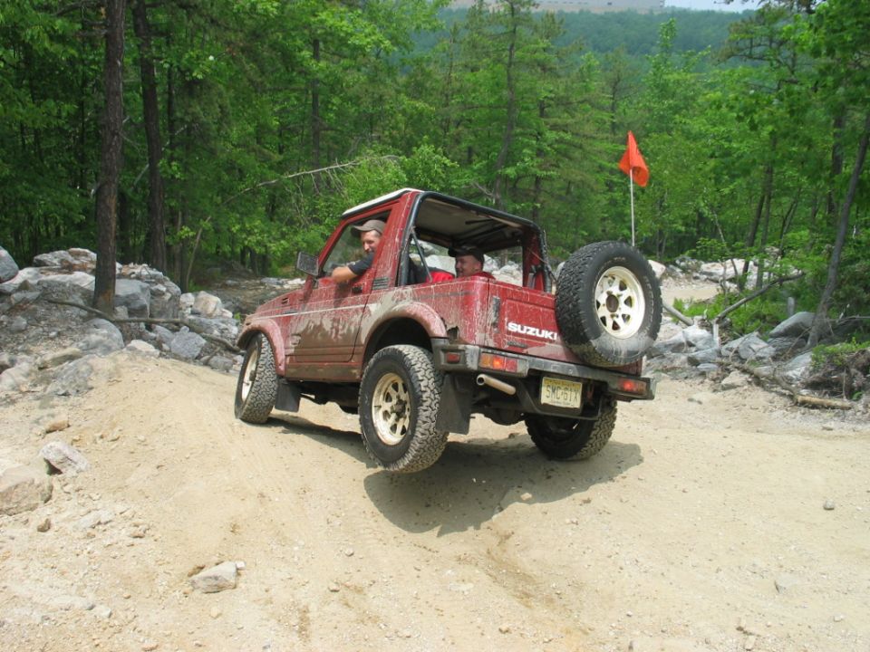 Muddy red Suzuki Samurai with two occupants climbing a rocky dirt trail
