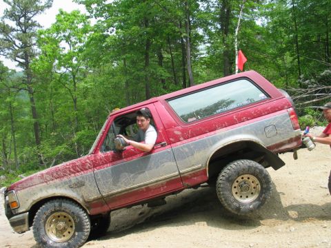 1995 Ford Bronco - Red and silver two-tone full-size SUV with a removable hardtop climbing a rocky dirt trail, driver's side view with the front wheel lifted…