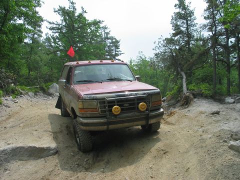 1995 Ford Bronco - Dusty red full-size Ford Bronco on a rutted rock and dirt trail, viewed head-on from slightly left. It carries a tubular brush guard with t…
