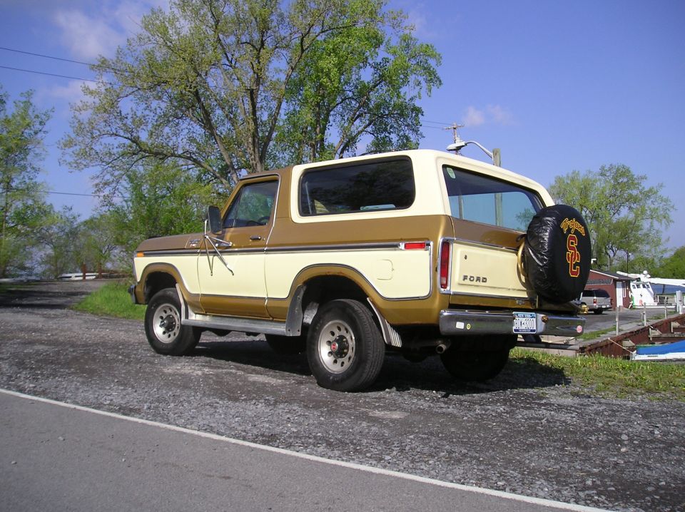 Rear 3/4 view of tan and brown Ford Bronco SUV with USC Trojans spare tire cover, parked on gravel.