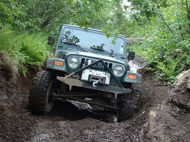 Green Jeep Wrangler on a muddy trail, front-left view, with aftermarket bumper and winch