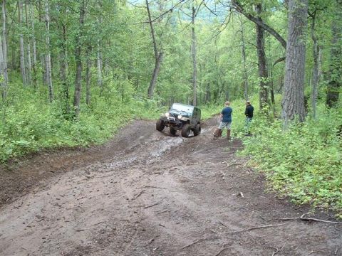 2000 Jeep TJ - A Jeep TJ navigates a challenging muddy path through a dense forest, with onlookers observing the off-road adventure.