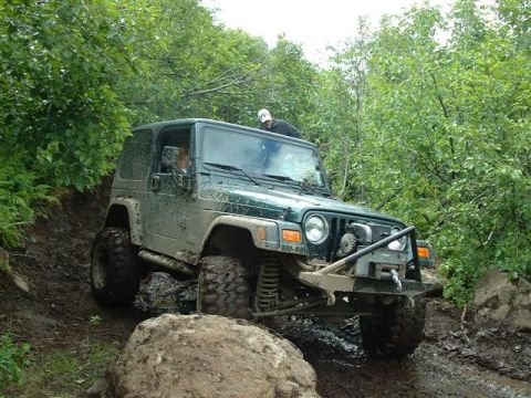 2000 Jeep TJ - A green 2000 Jeep TJ navigates a challenging, muddy off-road course surrounded by dense foliage.