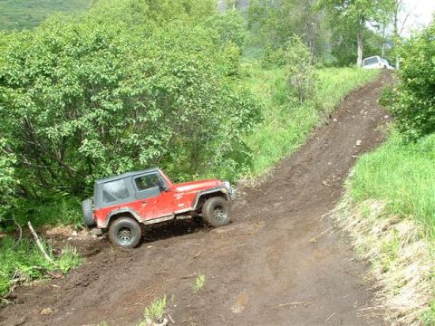 2000 Jeep TJ - A red Jeep TJ navigates a steep, muddy incline surrounded by lush green foliage.