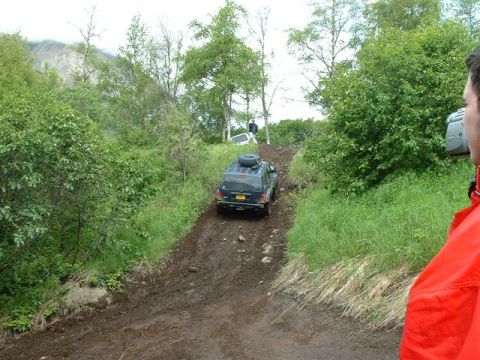 2000 Jeep Wrangler - A dark SUV navigates a steep, muddy trail with lush green foliage on either side.