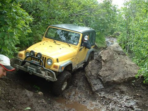 2000 Jeep Wrangler - A yellow 2000 Jeep TJ tackles a challenging muddy off-road course, maneuvering around large rocks.
