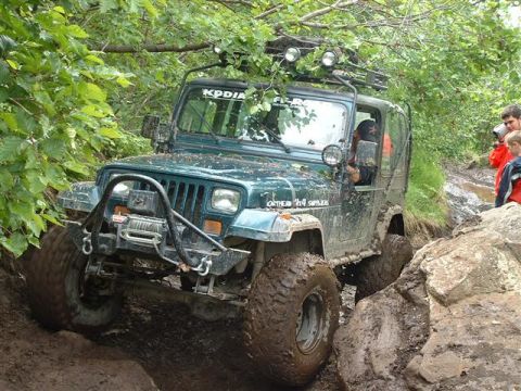 2000 Jeep Wrangler - A green Jeep Wrangler navigates a challenging muddy trail with large rocks.