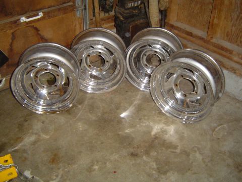 1989 Ford Bronco - A set of four chrome wheels are resting on a concrete floor in front of a wooden garage door.