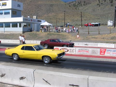 1972 Ford Mustang - Two classic muscle cars compete on a drag strip under a clear sky with mountains in the distance.