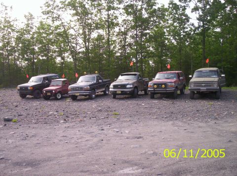 1995 Ford Bronco - Six lifted 4x4s lined up on a gravel staging lot at the edge of the woods, most flying orange whip flags: a black Bronco, a small red Suzuk…