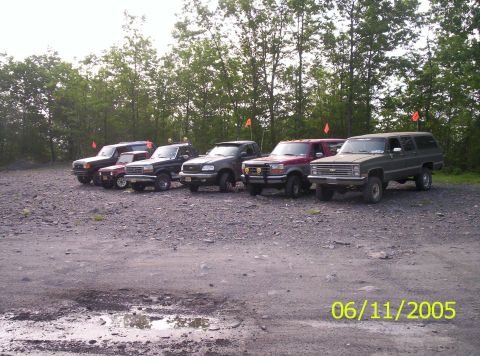 1995 Ford Bronco - A lineup of five off-road trucks and SUVs parked in a gravel staging lot, several flying tall orange trail flags; a green Chevrolet Suburba…