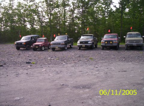 1995 Ford Bronco - A collection of trucks and SUVs are parked in a gravel lot on a cloudy day.