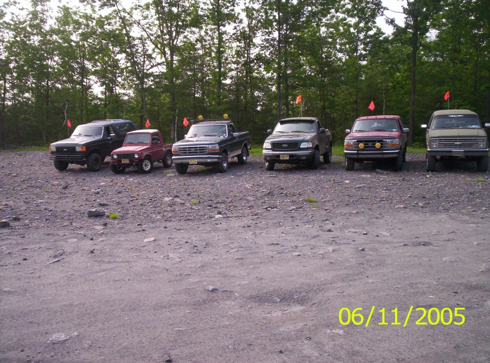 Several trucks and a small SUV parked on gravel lot with trees in background
