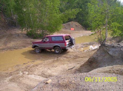 1995 Ford Bronco - Elevated side view of a maroon-over-gray full-size Ford Bronco wading through a muddy brown water hole in a sand-and-gravel pit, photograph…