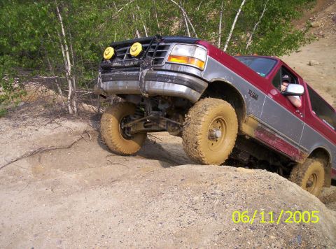 1995 Ford Bronco - Lifted two-tone red and silver full-size Ford Bronco climbing a rock ledge on a dirt trail, front driver-side tire lifted clear of the grou…
