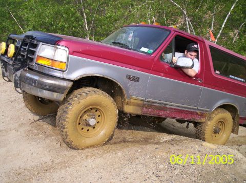 1995 Ford Bronco - A red and silver 1995 Ford Bronco navigates a muddy off-road trail, kicking up dirt.