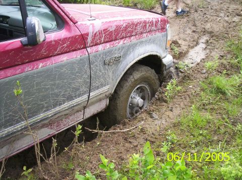 1995 Ford Bronco - Close side view of a red and silver two-tone Ford Bronco buried to the hub in a deep mud rut, with the front tire and alloy wheel packed so…