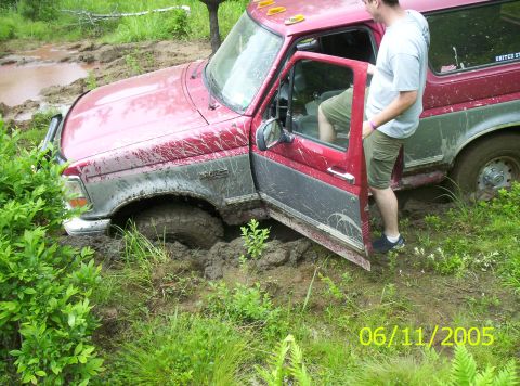 1995 Ford Bronco - Red-over-silver Ford Bronco buried to the axles in a mud hole with the driver's door hanging open and a man standing in the muck alongside.…
