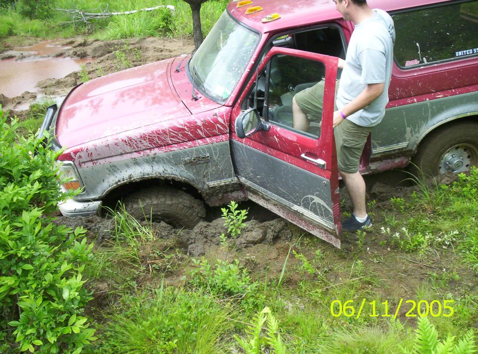 Red and silver Ford Bronco stuck axle-deep in mud with driver door open