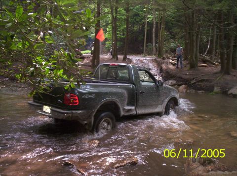 1995 Ford Bronco - Dark green regular-cab Ford F-150 with a 4x4 bedside decal drives through a muddy forest creek, water washing over the tires and mud spatte…