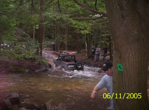 1995 Ford Bronco - A 1995 Ford Bronco navigates a stream in a wooded area, with onlookers present.